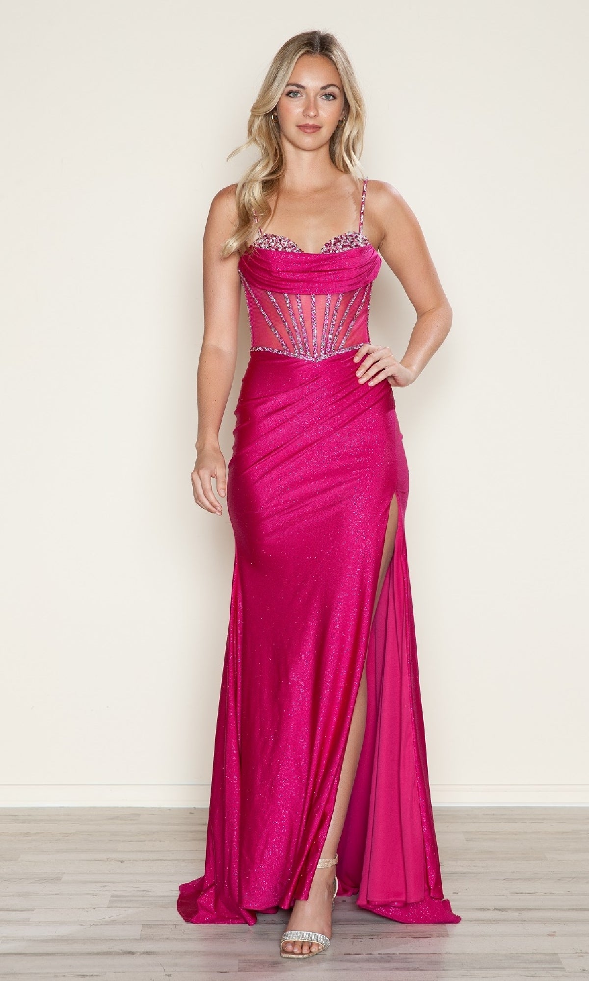 A woman stands on a light wood floor in the Sheer Beaded-Corset Long Prom Dress: Poly USA 9750—a fitted, bright pink gown with spaghetti straps, a beaded sheer corset bodice, and a high leg slit—against a plain background.